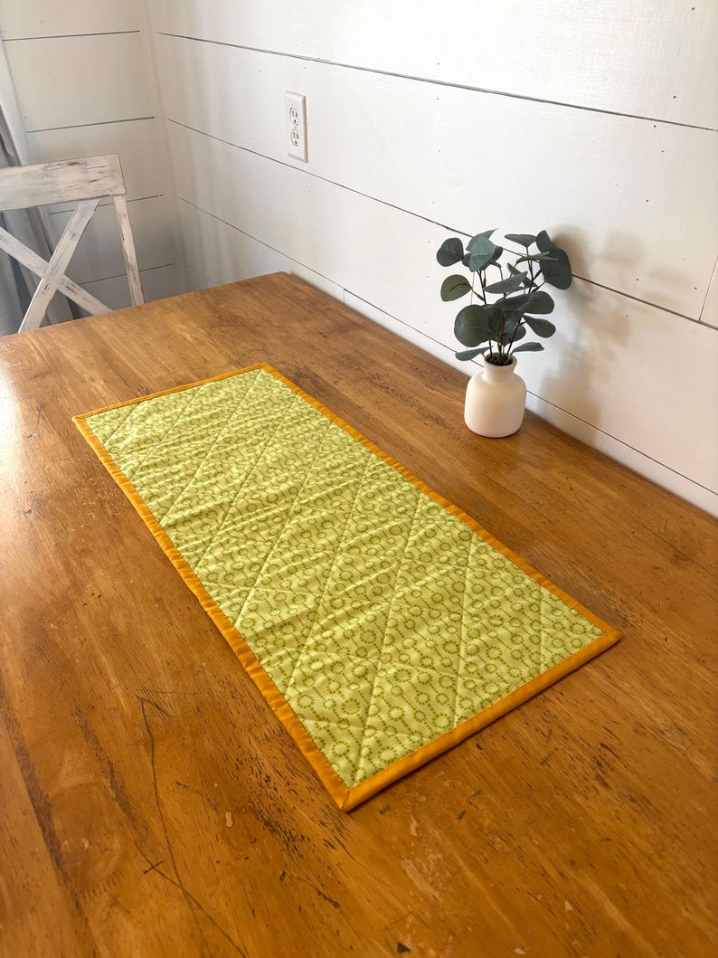 May include: A rectangular, quilted table runner with a light green floral pattern and a gold border. The runner is placed on a wooden table. A small white vase with greenery is in the background.