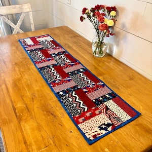May include: A rectangular fabric table runner featuring a patchwork design of red, white, and blue patterns, including stars, stripes, and zigzags. The runner is finished with a solid blue border and is laid flat on a wooden surface.