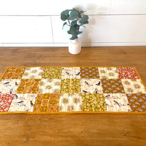 May include: A quilted table runner with a patchwork design. The runner features squares of various patterns, including birds, floral prints, and sunbursts, in shades of gold, green, and red. The edges are finished with a yellow border.