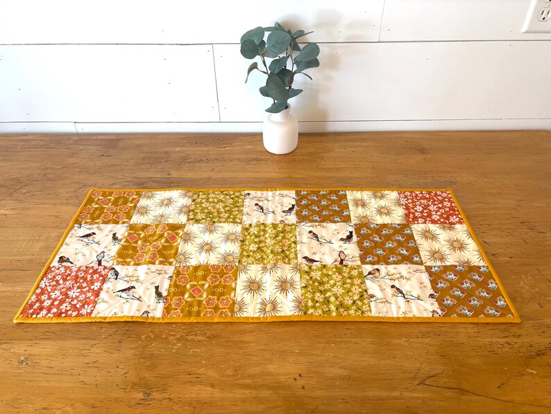 May include: A quilted table runner with a patchwork design. The runner features squares of various patterns, including birds, floral prints, and sunbursts, in shades of gold, green, and red. The edges are finished with a yellow border.