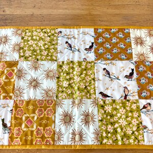 May include: A quilted table runner with a patchwork design. The squares feature floral, sunburst, and bird patterns in shades of gold, white, and green. The runner is trimmed with a gold border and has a quilted texture.