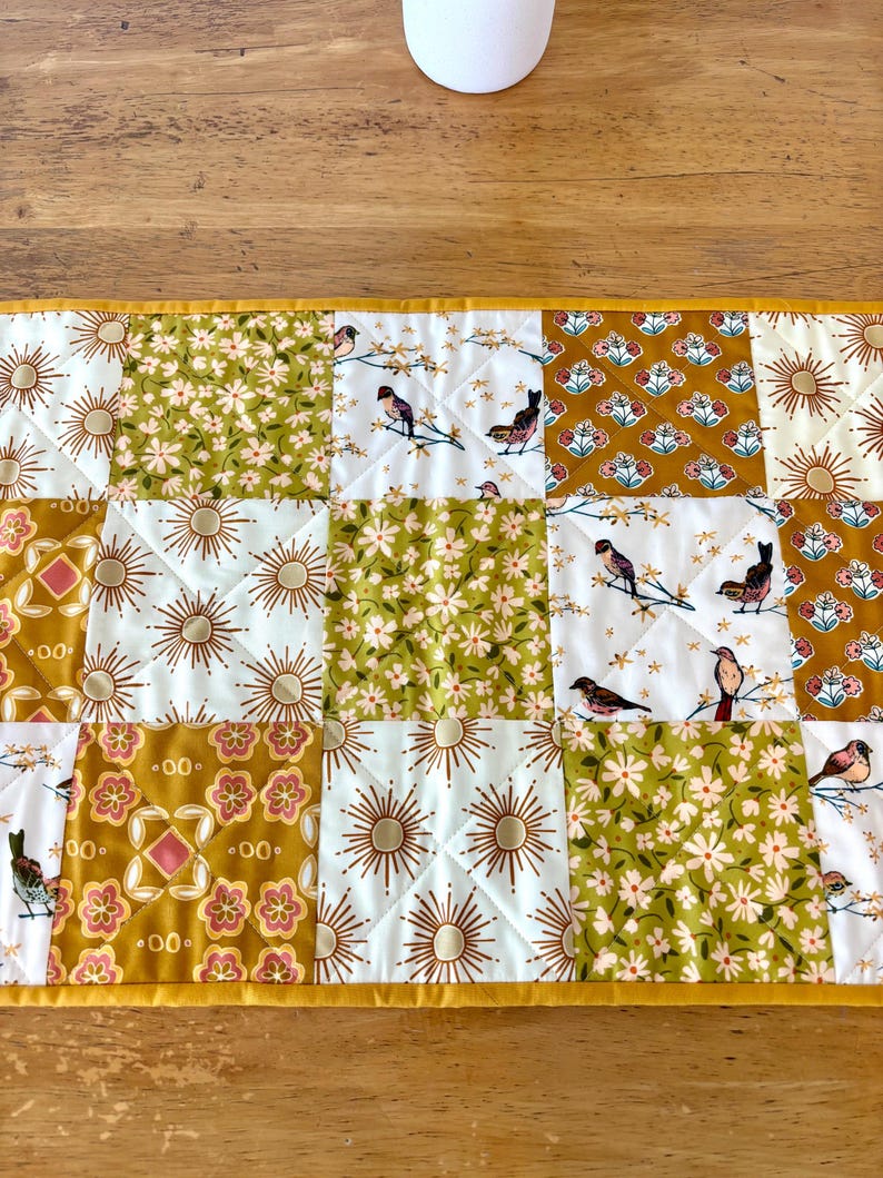 May include: A quilted table runner with a patchwork design. The squares feature floral, sunburst, and bird patterns in shades of gold, white, and green. The runner is trimmed with a gold border and has a quilted texture.
