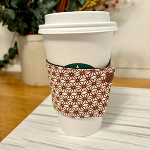 May include: A white coffee cup with a brown and white patterned fabric sleeve. The sleeve has a brown button on the side.