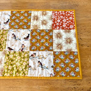 May include: A quilted table runner with a patchwork design. The squares feature various patterns, including birds on branches, floral prints, and sunbursts, in shades of gold, white, and red. The runner is bordered with a solid yellow fabric.