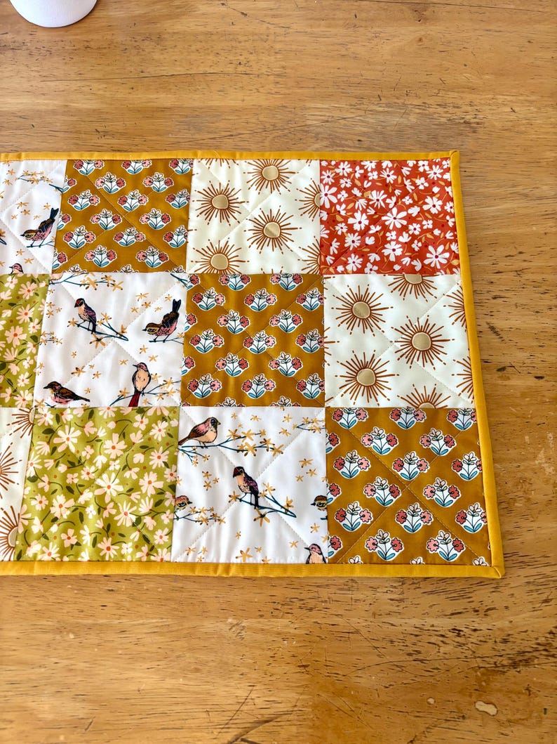 May include: A quilted table runner with a patchwork design. The squares feature various patterns, including birds on branches, floral prints, and sunbursts, in shades of gold, white, and red. The runner is bordered with a solid yellow fabric.