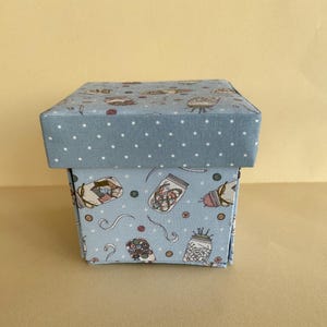 May include: A square gift box with a light blue lid and sides. The lid has white polka dots. The sides of the box are decorated with illustrations of mason jars filled with various items, along with other decorative elements.