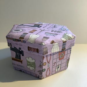 May include: A hexagonal sewing box with a lavender background and a repeating pattern of sewing tools. The box features images of sewing machines, spools of thread, scissors, and thimbles. The box is made of fabric.