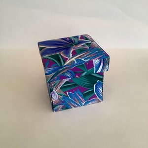 May include: A square gift box with a lid, decorated with a colourful floral pattern. The design features blue, purple, and green leaves and flowers on a dark background. The box is set against a plain white background.