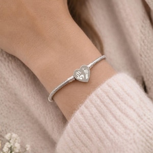 May include: A silver-toned snake chain bracelet featuring a central heart-shaped charm. The charm is engraved with a detailed white graphic of a small handprint and a footprint, creating a clear contrast against the polished metallic surface.