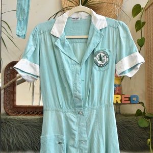 Puede incluir: Un vestido turquesa vintage con cuello y puños blancos, colgado de una percha de madera. El vestido presenta una parte delantera abotonada, mangas cortas y un parche en el pecho. Un cinturón a juego está colgado cerca.