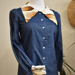 May include: A blue denim shirt with a large, patterned collar and cuffs. The collar and cuffs are made of a white fabric with brown and beige abstract shapes.