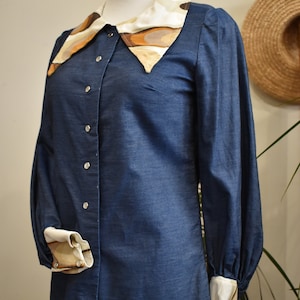 May include: A blue denim shirt with a large, patterned collar. The collar is made of a white fabric with brown and beige abstract shapes. The shirt has long sleeves with white cuffs and button closures.