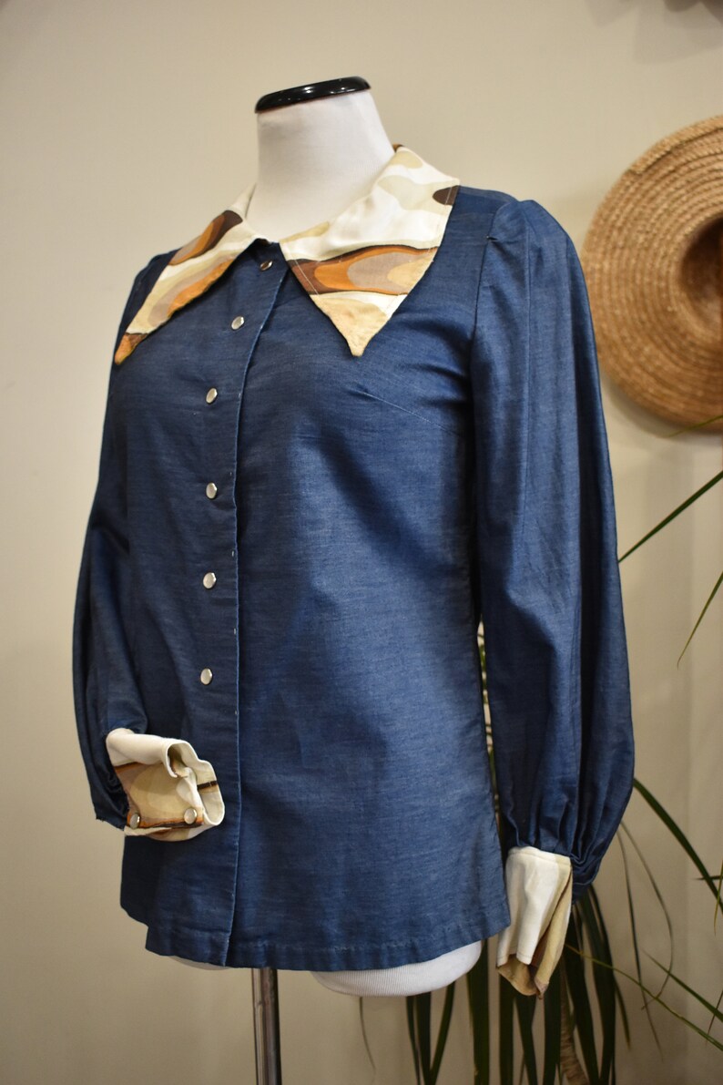 May include: A blue denim shirt with a large, patterned collar. The collar is made of a white fabric with brown and beige abstract shapes. The shirt has long sleeves with white cuffs and button closures.