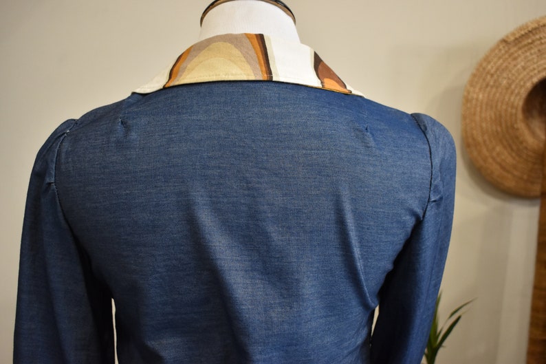 May include: A blue denim shirt with a long sleeve and a patterned collar. The collar is made of a brown and beige fabric with a geometric design.