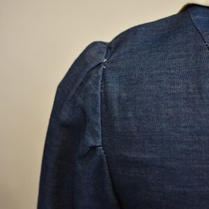 May include: Close-up of the shoulder seam of a dark blue denim jacket with a gathered puff sleeve.