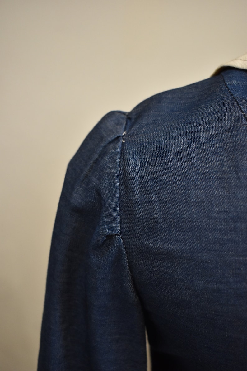 May include: Close-up of the shoulder seam of a dark blue denim jacket with a gathered puff sleeve.