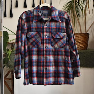 May include: A long-sleeved, button-down shirt in a plaid pattern of red, blue, gray, and black. The shirt has two chest pockets and a collar. The shirt is hanging on a white hanger.