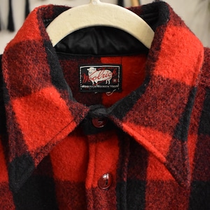 May include: A close-up of a red and black plaid wool jacket. The jacket has a black collar and a black satin lining. The jacket has three buttons and a label that reads "Woolrich Woolen Mills".