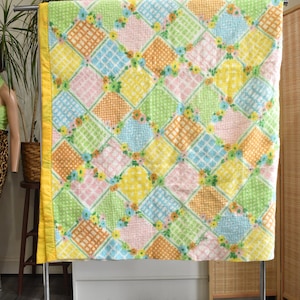 Op de afbeelding: Een geel, roze, groen en blauw patchwork-deken met een bloemenpatroon. Het deken hangt aan een metalen rek met wielen.