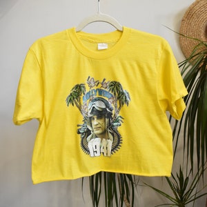 Può includere: Maglietta gialla a maniche corte e crop con una stampa grafica di un uomo con un casco da pilota e occhiali. La grafica include anche palme, pistole e il testo "Hollywood 1941".