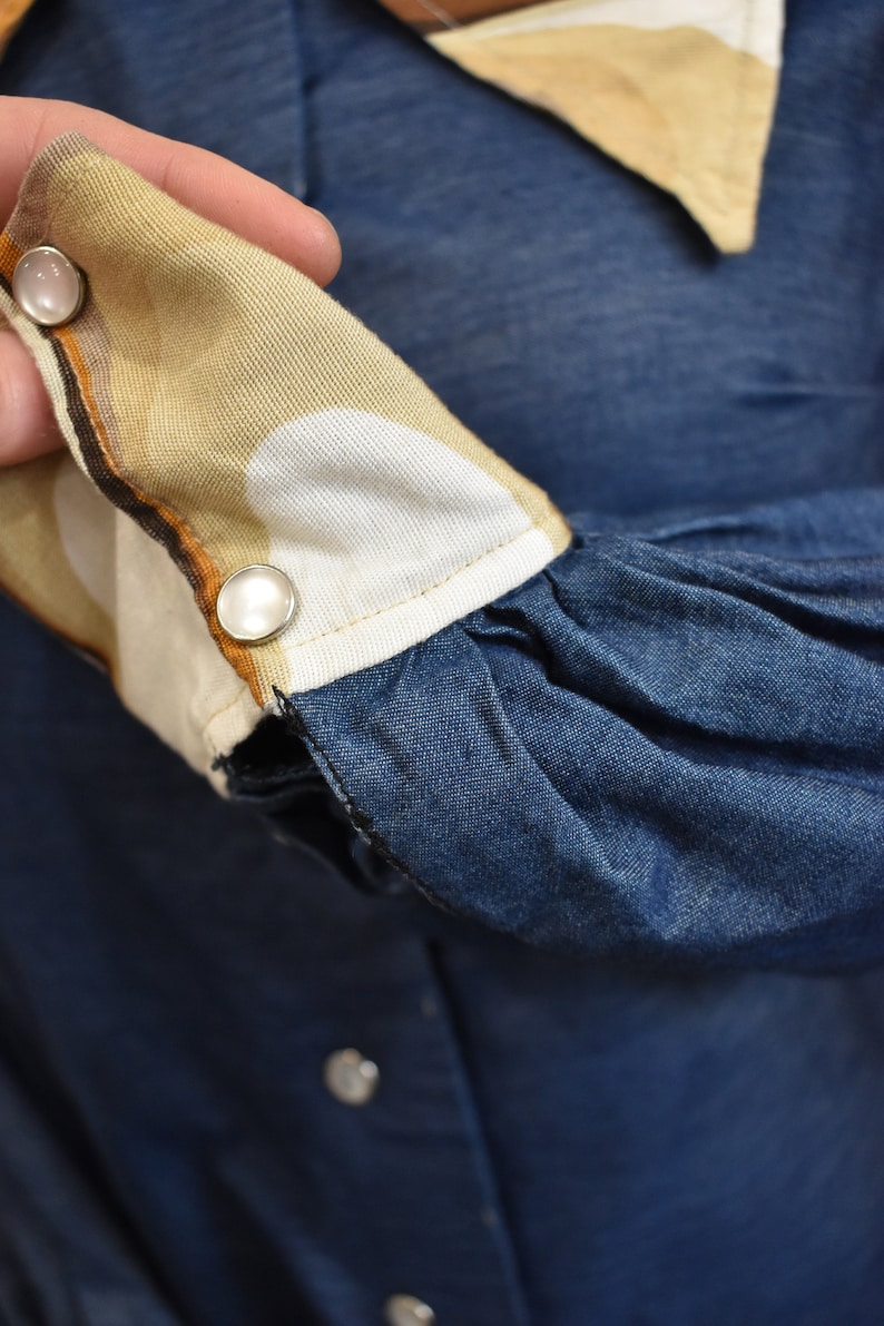 May include: Close-up of a denim shirt with a brown and white patterned cuff. The cuff has two white buttons.