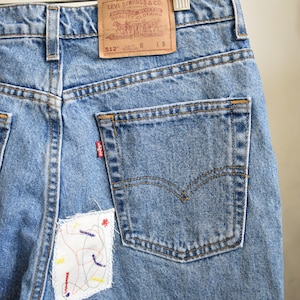 May include: Light blue denim jeans with a back pocket and a small embroidered patch. The jeans have a Levi's label with the text "Levi Strauss & Co. San Francisco, Cal. Original Quality Clothing" and "512" printed on it.