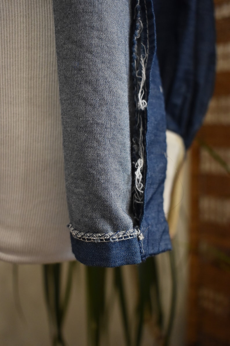 May include: Close-up of the seam of a blue denim shirt. The seam is frayed and has a white thread running through it.