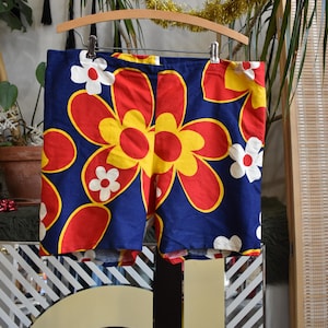 Può includere: Shorts blu con una grande stampa floreale in rosso, giallo e bianco. I fiori sono stilizzati e assomigliano a margherite.