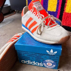 Peut inclure: Baskets Adidas Nite Jogger crème avec des rayures et des accents orange, posées sur une boîte Adidas bleue. Les baskets ont un design rétro avec une semelle bleue et une semelle extérieure en caoutchouc marron. La boîte porte le logo Adidas.