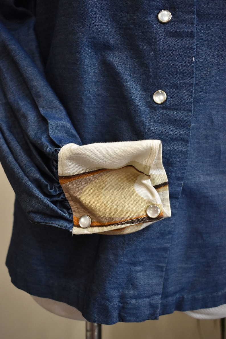 May include: A blue denim shirt with a white and brown patterned cuff. The shirt has three white buttons.