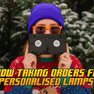 A person wearing a purple beanie and orange sunglasses holds a black VHS tape in front of their face, with snow-covered pine trees in the background.
