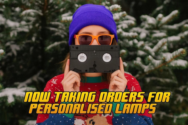 A person wearing a purple beanie and orange sunglasses holds a black VHS tape in front of their face, with snow-covered pine trees in the background.