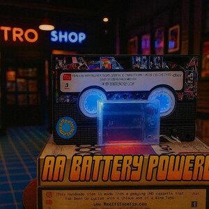 a retro-style cassette tape and a battery-powered cassette tape, both displayed in a dimly lit room with neon signs in the background.