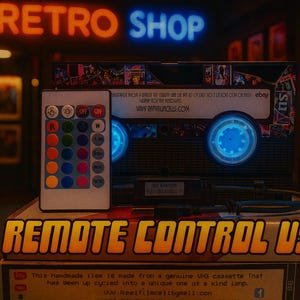 a remote control USB device, which is a handmade item made from a genuine VU cassette tape. The device has been upcycled into a unique one at a kind lamp. The remote control USB is displayed on top of a stack of books, with a neon sign reading "RETRO SHOP" in the background.