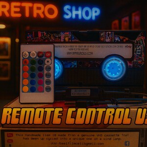 a remote control USB device, which is a handmade item made from a genuine VU cassette tape. The device has been upcycled into a unique one at a kind lamp. The remote control USB is displayed on top of a stack of books, with a neon sign reading "RETRO SHOP" in the background.
