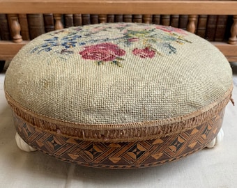 May include: A round, beige, needlepoint footstool with a floral design. The footstool has a wooden base with a geometric pattern and white wooden feet.