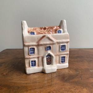 May include: A cream-coloured ceramic house ornament with blue window panes and a brown roof. The front door is dark brown. The house has a classic design, likely a decorative item, and is reminiscent of a traditional home.