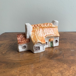 May include: A ceramic cottage figurine with a thatched roof, white walls, and small windows. The roof is a warm orange color, and the windows have a crosshatch design. The cottage has a small green door and a chimney.