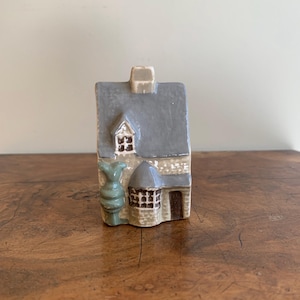 May include: A miniature ceramic house with a gray roof, beige chimney, and a small green decorative element. The house has a stone-like texture and a brown door. The house is on a wooden surface.