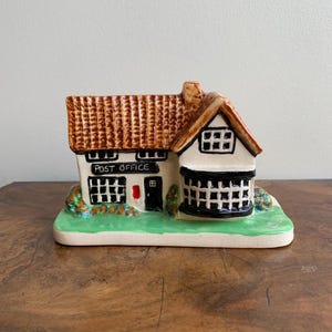 May include: A ceramic miniature model of a post office building with a brown roof, white walls, and black trim. The building has a sign that reads "POST OFFICE" and a small red door.