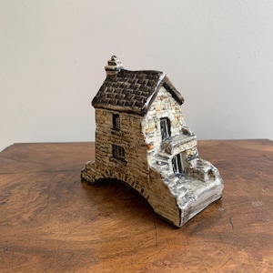 May include: A ceramic miniature cottage with a stone facade, dark brown roof, and small windows. The cottage has a bridge-like base and a chimney. The color palette includes beige, brown, and black.