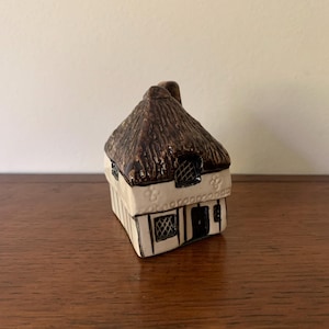 May include: A miniature ceramic cottage figurine featuring a dark brown textured thatched roof, cream-colored walls with black timber framing, and small lattice-patterned windows. The structure includes a central dark door and decorative floral accents.