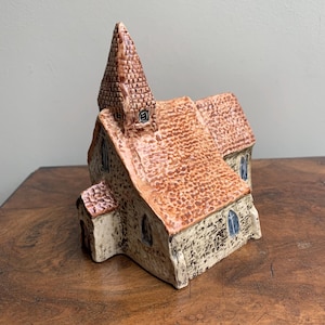 May include: A ceramic church model with a textured, reddish-brown roof and a tall steeple. The walls are a light beige with small arched windows. The model is placed on a wooden surface.