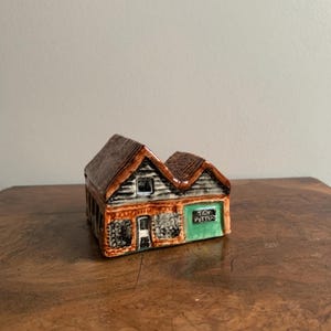 May include: A small ceramic model of a house with a brown roof, gray stone walls, and a green door. The door has the words "Tea Potters" written on it.