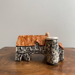 May include: A ceramic miniature of a stone archway with a red-tiled roof and a cylindrical tower. The archway is painted in shades of gray and black, with small windows and a textured appearance. The tower has a white top.