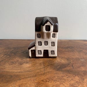 May include: A small ceramic house figurine with a white body and brown roof and trim. The house has multiple windows and a door.