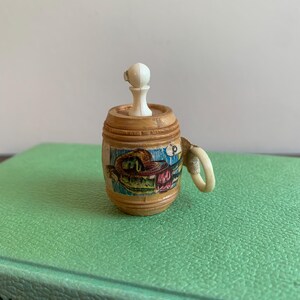 May include: A miniature wooden beer stein with a handle and a white stopper. The stein is cylindrical with horizontal grooves and a colorful illustration around the middle. The stein is resting on a green surface.