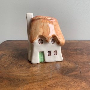 May include: A ceramic cottage ornament with a thatched roof and a chimney. The cottage is painted white with a tan roof and a green door. The windows are small and have a brown interior. The ornament is on a wooden surface.