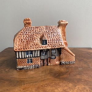 May include: A ceramic cottage ornament with a brown tiled roof, brick walls, and two chimneys. The cottage features blue window frames and black and white timber framing. The ornament is set on a wooden surface.