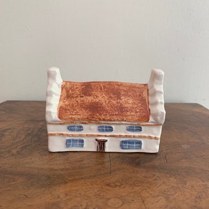 May include: A small white ceramic house with a brown roof and blue windows. The house has a brown door and is sitting on a wooden surface.